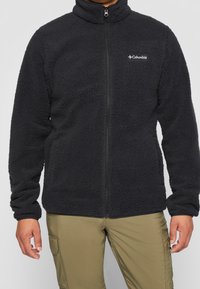 Giacca in pile nera con colletto alto, zip frontale e tasche laterali; presenta una superficie texturizzata e un piccolo logo sul petto.