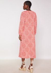 Vrouw met kort haar, gekleed in een lange rood-witte jurk met patroon en beige espadrilles met hak en enkeltouwen, met de rug naar ons gekeerd.