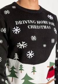 Maglione nero a maglia con la scritta "TORNANDO A CASA PER NATALE", fiocchi di neve bianchi, alberi verdi e un disegno di un'auto rossa. Tessuto morbido.
