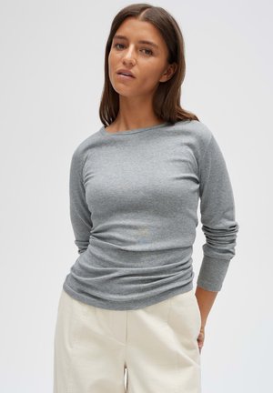 Frau mit glattem braunem Haar, die ein graues Langarmshirt und hellbeige Hose trägt, steht vor einem schlichten hellen Hintergrund.
