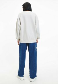 Sudadera de gran tamaño en gris claro con hombros caídos y puños acanalados, combinada con pantalones de denim azul sueltos que tienen un parche de logo en el lateral.