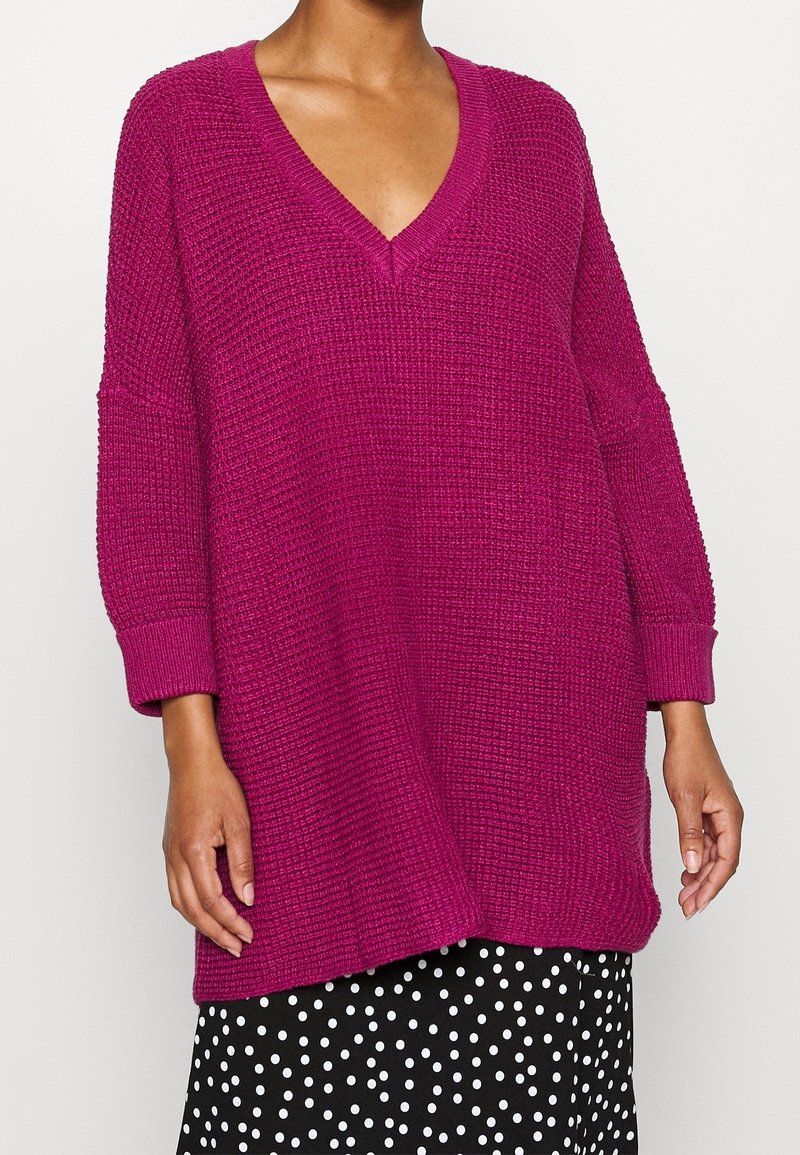 Pull surdimensionné en maille de couleur magenta vif avec un col en V et des manches 3/4. Tissu texturé, avec une coupe décontractée et des poignets côtelés.