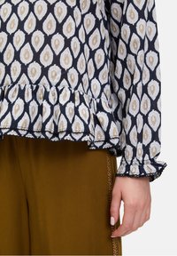 Granatowa bluzka w biało-złoty wzór paisley, z falbaniastym wykończeniem u dołu i na mankietach, z długimi rękawami, wykonana z lekkiego materiału. Połączona z brązowymi spodniami.