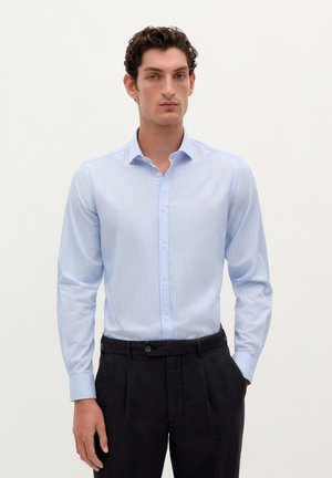 Cămașă albastru deschis cu dungi verticale albe, cu mâneci lungi și guler clasic. Stilizată cu pantaloni închiși la culoare.