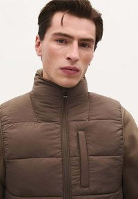 Braune wattierte Weste mit hohem Kragen, Reißverschluss und vertikalen Taschendetails. Das Material wirkt glatt mit einer gesteppten Textur.