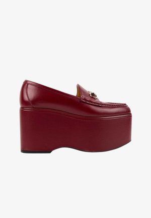 Mocassin à plateforme en cuir rouge foncé avec une semelle épaisse et un détail de boucle en métal sur le dessus, présenté de côté.
