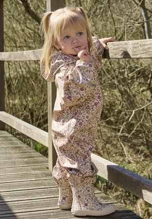 Tuta impermeabile con stampa floreale color crema con motivi rosa e viola, abbinata a stivali di gomma beige, in piedi su un ponte di legno.