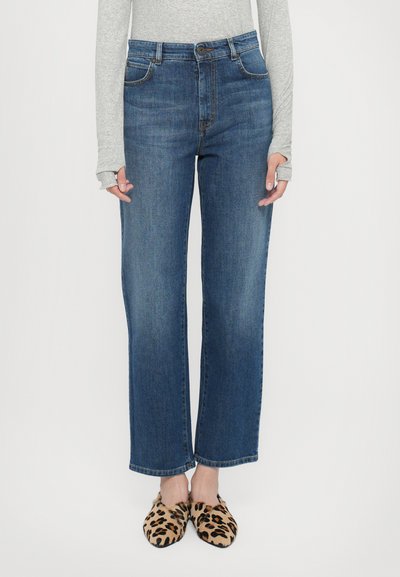 Blaue Jeans mit hoher Taille und geradem Bein. Verfügt über Fronttaschen und einen Knopfverschluss. Getragen mit leichtem Leopardenmuster bei Slippers.