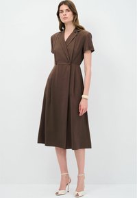 Robe enveloppante marron à manches courtes avec un col, une jupe plissée et des boutons à la taille, accompagnée de sandales à talons de couleur claire.