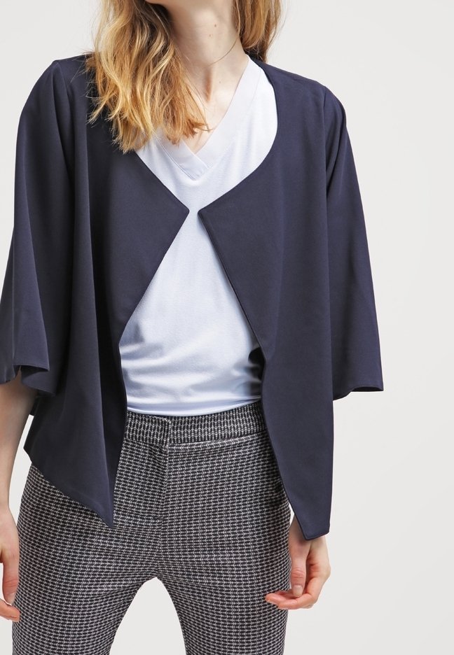 Personne portant une veste bleu marine ouverte devant, un t-shirt blanc à col en V et un pantalon à carreaux noir et blanc, montrée du cou aux hanches.