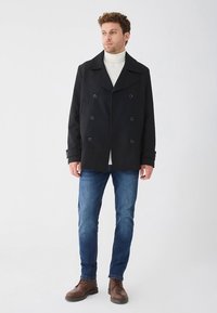 Czarny dwurzędowy płaszcz peacoat z szerokimi klapami i dużymi guzikami, noszony na białym golfie, zestawiony z niebieskimi dżinsami i brązowymi butami.