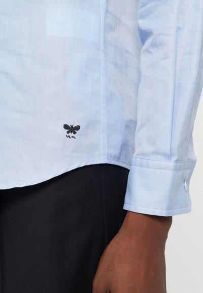 Lys blå skjorte lavet af et letvægtsstof, med et sort broderet bi-logo nær kanten og en manchet-sleeve design.