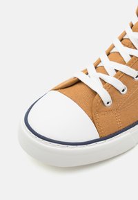 Brune kanvas sneaker med hvit gummitå, marineblå detaljer og hvite snøringer. Har strukturert stoff og forsterkede sømmer.