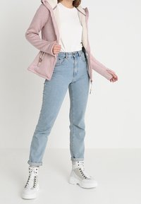 Giacca foderata in pile rosa con cappuccio, abbinata a jeans in denim blu chiaro e sneakers alte bianche con accenti metallici.