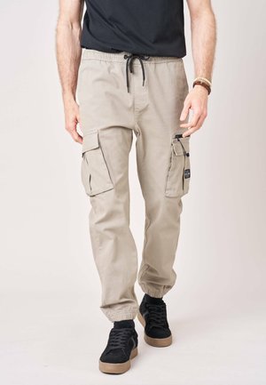 Man draagt beige cargobroek met elastische boorden, een zwarte taille met trekkoord en zwarte sneakers met bruine zolen.