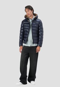 Giacca puffer navy con cappuccio e zip, caratterizzata da un design trapuntato e una foderatura interna marrone. Indossata con una camicia leggera e jeans scuri.