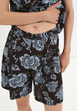 Person trägt dunkle Shorts mit großem hellblauem Blumenmuster, hält ein passendes Oberteil hoch, um den Bauch und den Bauchnabel zu zeigen.