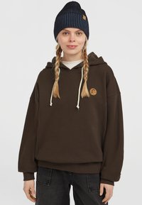 Bruine hooded sweatshirt met trekkoord, geribbelde manchetten en een cirkelvormig logo. Gekoeld met een marineblauwe gestreepte beanie.
