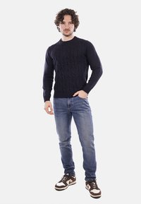 Uomo in piedi con una mano in tasca, indossa un maglione scuro lavorato a trecce, jeans blu e sneakers marroni e bianche.