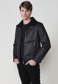 Giacca in pelle nera con un finish testurizzato, dotata di colletto in shearling nero, chiusura con zip e tasche laterali. Indossata sopra una camicia nera.