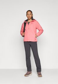 Vrouw in een roze hoodie, met donkere grijze broek en bruine wandelschoenen, staand met één hand in de zak en een rugzakriem over haar schouder.