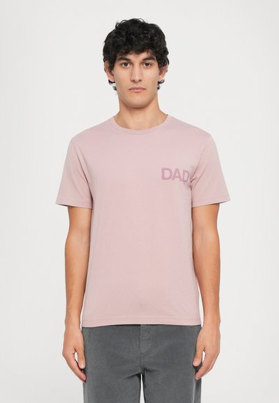 Camiseta de manga corta, de color rosa claro, hecha de una tela suave, con un sutil gráfico de "DAD" en la parte izquierda del pecho. Corte casual, cuello redondo.
