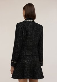 Donna con i capelli corti castani che indossa una giacca nera a trama e una gonna svasata coordinata, vista di schiena su uno sfondo semplice.