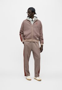 Μπουφάν με φερμουάρ σε σχέδιο houndstooth σε κόκκινο και μπεζ με κουκούλα· αντίστοιχο παντελόνι με κάθετες ρίγες στα πλάγια· φορεμένα με καπέλο με ρίγες και αθλητικά παπούτσια.