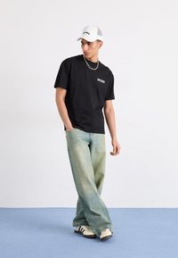 Camiseta de algodón negra con un pequeño logo blanco, combinada con unos vaqueros de pierna ancha de color azul claro. Una gorra de malla blanca y unas zapatillas en blanco y negro completan el look.