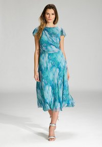 Sukienka o luźnym kroju, sięgająca kostek, w niebiesko-zielony abstrakcyjny wzór, z krótkimi falbanistymi rękawami i dopasowanym pasem. Nosi się ją z srebrnymi sandałkami na obcasie.