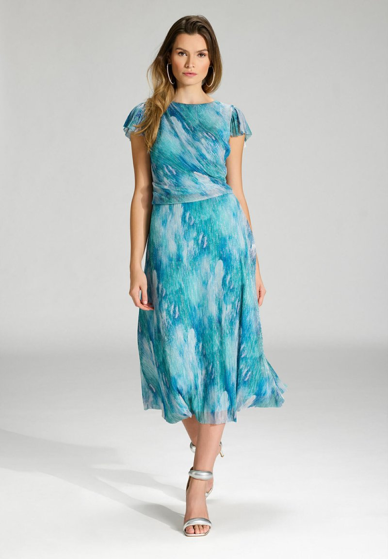 Sukienka o luźnym kroju, sięgająca kostek, w niebiesko-zielony abstrakcyjny wzór, z krótkimi falbanistymi rękawami i dopasowanym pasem. Nosi się ją z srebrnymi sandałkami na obcasie.