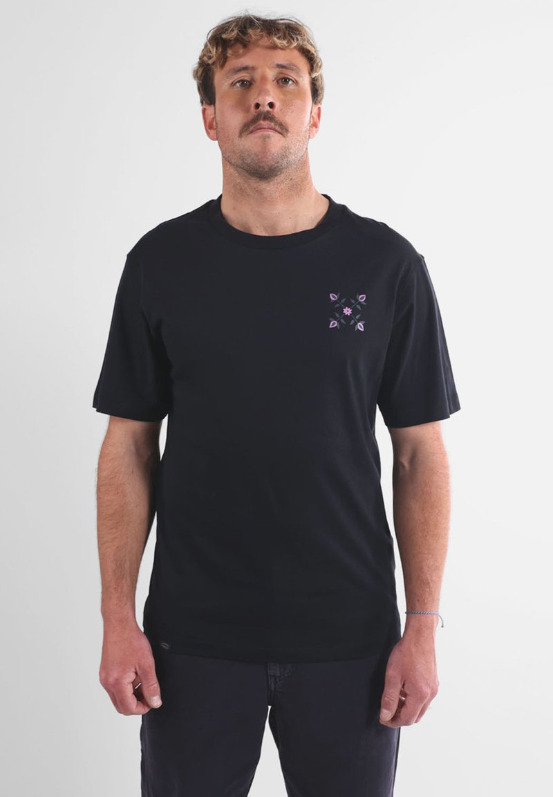 Homme aux cheveux brun clair et à la moustache, portant un t-shirt noir avec un petit motif floral violet sur le côté gauche de la poitrine, se tenant devant un fond blanc.