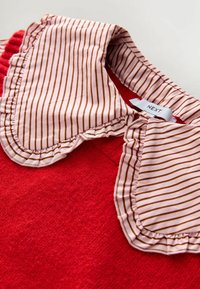 Rød trøje med en lyserød og brunstribet krave med flæsekant; synligt mærke med teksten "NEXT." Blød tekstur, nærbillede.