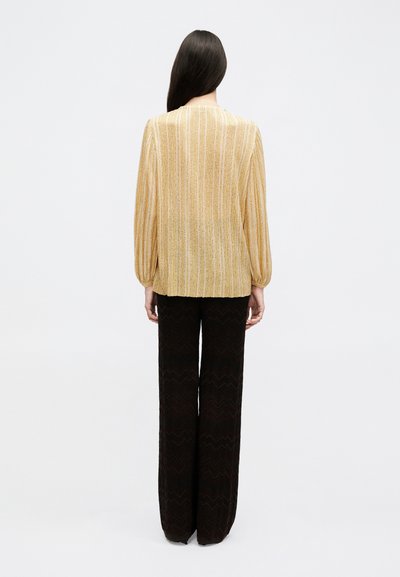 Γυναίκα που στέκεται γυρισμένη ανάποδα, φοράει μπλούζα με χρυσές ρίγες μακρυμάνικη και φαρδιά μαύρη παντελόνα, μπροστά σε απλό λευκό φόντο.