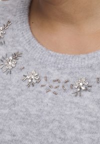 Pull en tricot gris orné de motifs floraux avec des cristaux clairs et des accents métalliques le long de l'encolure. Texture douce.