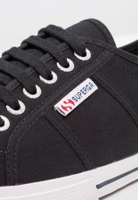 Černá plátěná teniska se stříbrnými průchodkami, kulatými tkaničkami a bílou gumovou podrážkou. Obsahuje značkovou štítek s logem v červené a modré barvě.