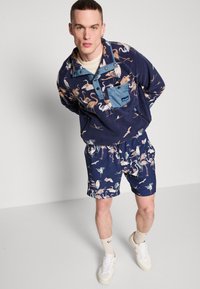 Fleece genser med en blå lomme og flamingomønster; kombinert med matchende shorts med et lignende mønster. Hvite joggesko og sokker.