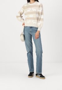 Maglione lavorato a maglia beige con motivo di renne bianche e fiocchi di neve, abbinato a jeans azzurri chiari e sneaker nere con strisce bianche.