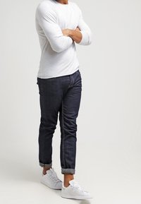 En man står med armarna i kors, iförd en vit långärmad skjorta, mörka uppkavlade jeans och vita sneakers framför en enfärgad bakgrund.