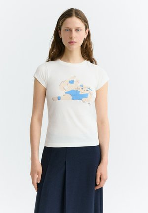 Femme aux longs cheveux bruns portant un t-shirt blanc avec un chat de dessin animé en vêtements de sport bleus et une jupe bleu marine sur un fond uni.