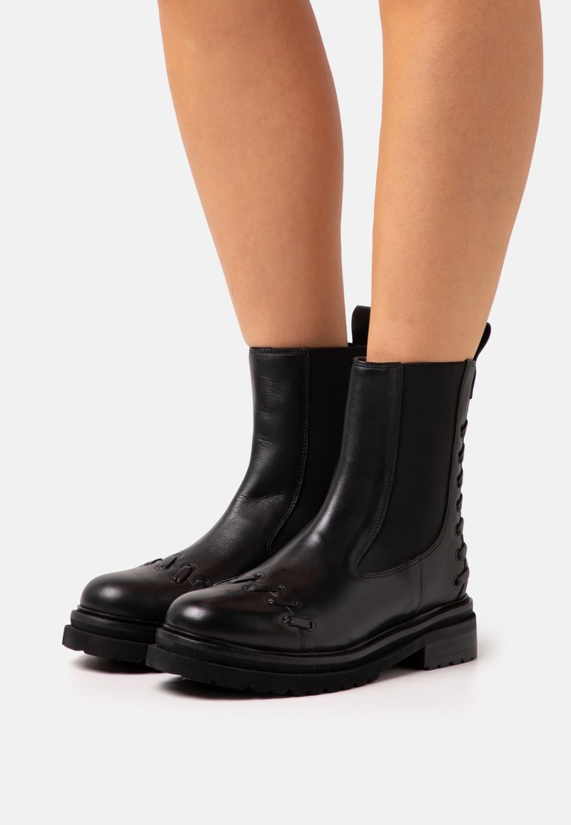 Mustat nahkaiset nilkkasaappaat, joissa on joustavat sivupaneelit, edessä nyöritettävä muotoilu ja paksut kumipohjat. Silkkinen pinta ja minimalistinen tyyli.