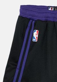 Calções de basquetebol pretos com uma cintura elástica roxa, apresentando textura de rede e um emblema do logótipo da NBA na lateral. Duas riscas roxas ao lado.