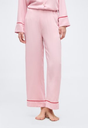 Γυναίκα που φοράει ανοιχτό ροζ σατέν παντελόνι πιτζάμας με κόκκινη περίγραμμα κοντά στις μανσέτες, όρθια ξυπόλυτη μπροστά σε απλό φόντο.