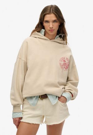 Sudadera oversized beige con un logo rosa, superpuesta sobre una camisa de rayas azul claro, combinada con pantalones cortos blancos deshilachados. Textura suave, ajuste casual.
