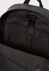 Čierny batoh zvnútra vyrobený z nylonovej tkaniny, obsahujúci zipsovú kapsu, štítok a slučku na zabezpečenie predmetov. Hladká textúra po celom povrchu.