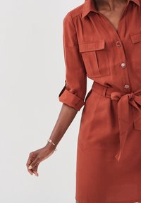 Robe chemise couleur rouille avec manches retroussables, deux poches poitrine, devant boutonné et taille nouée, présentant une texture lisse et un design structuré.