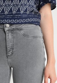 Jeans de mezclilla gris con una forma ajustada, que cuentan con bolsillos delanteros y cierre de botón, combinados con una blusa de patrón azul marino.