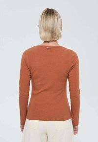 Haut à manches longues couleur rouille en texture côtelée avec un col ras du cou et un subtil logo au dos. Silhouette ajustée, tissu lisse.