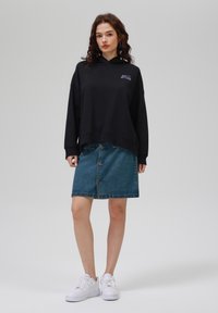 Sudadera negra con un bolsillo frontal, combinada con una falda de mezclilla azul clara que tiene botones en la parte delantera. Zapatillas blancas completan el look.