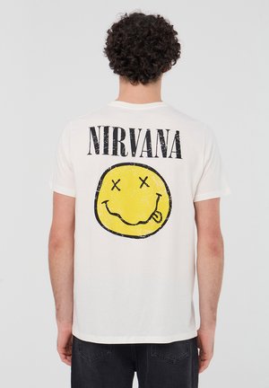 T-shirt bianco in cotone con la scritta nera "NIRVANA" in grande sopra un grafico di una faccina sorridente gialla con gli occhi incrociati e la lingua di fuori.